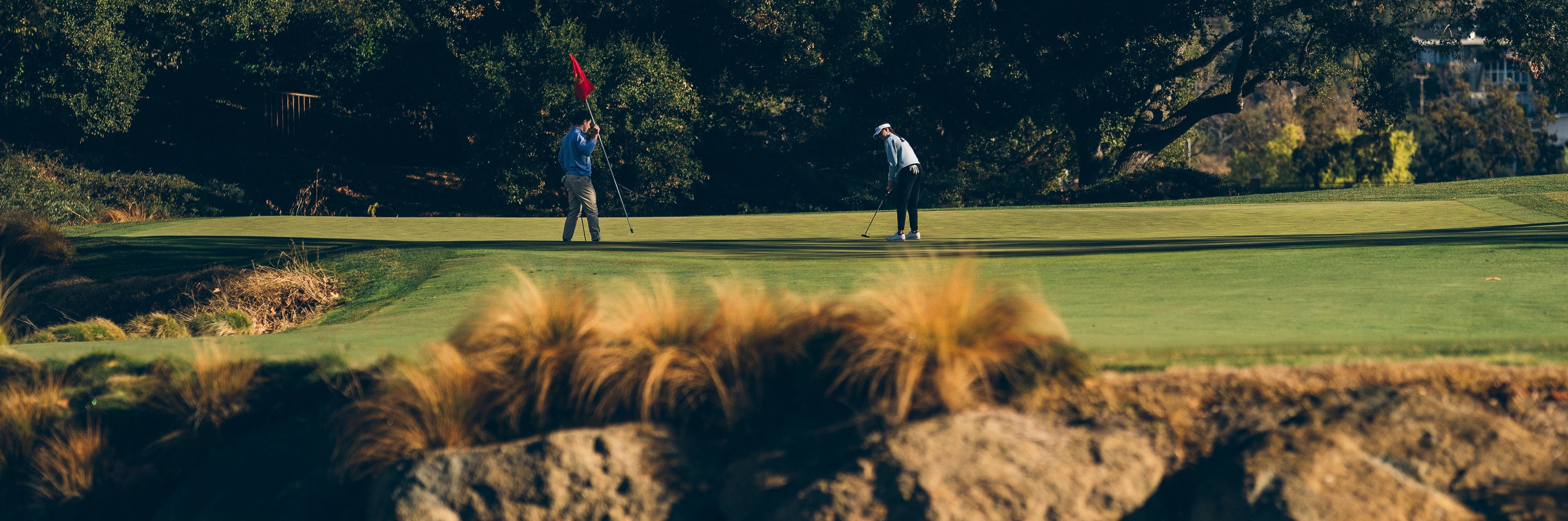 Two golfers on a green, one putting and another holding the flagstick.