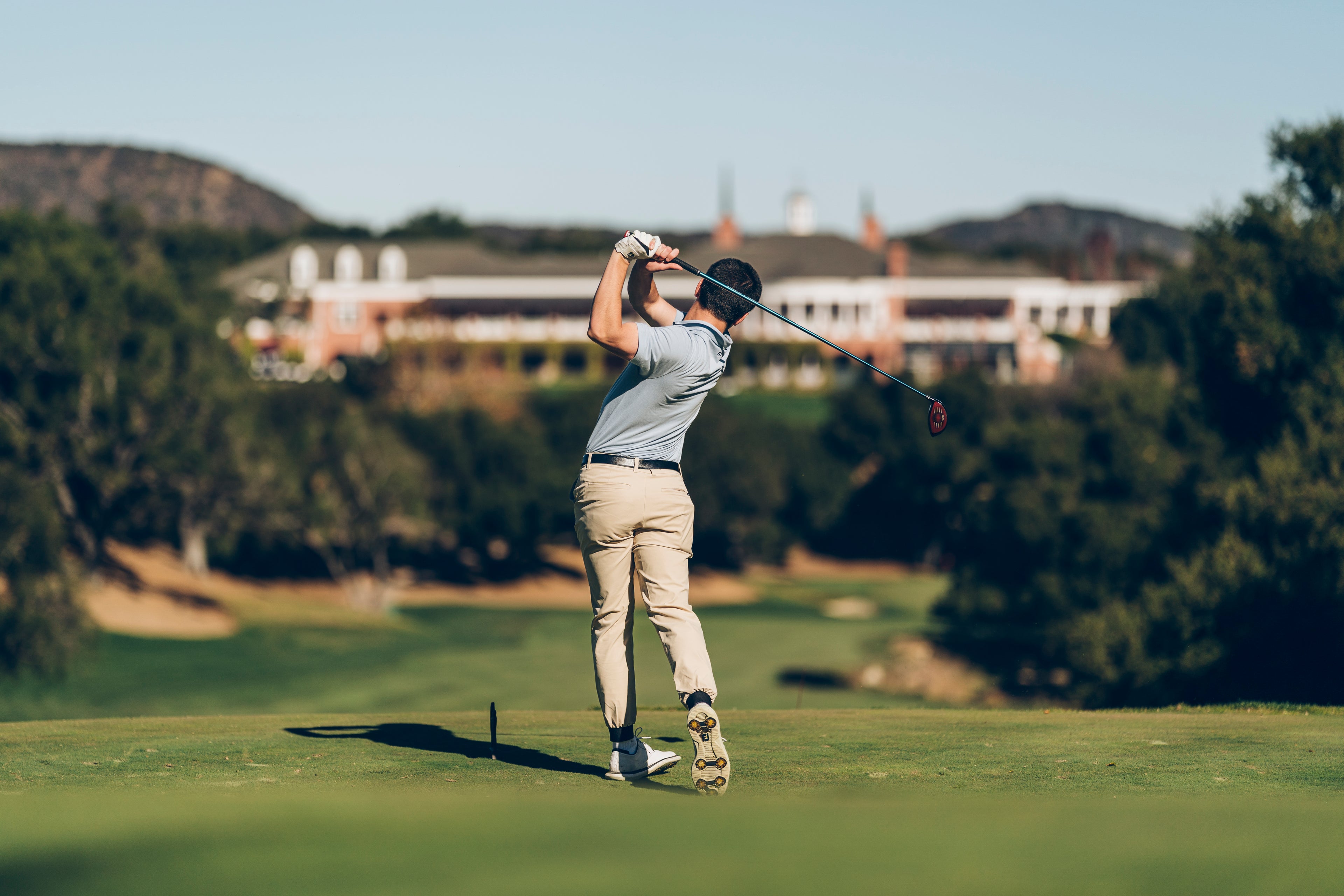A person playing golf on a lush green course swings a club toward a distant building.