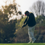 A person wearing a cap and jacket is swinging a golf club on a grassy course.