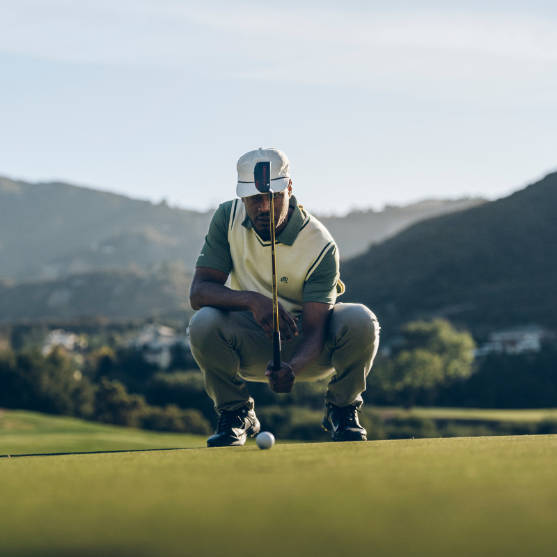 A golfer crouches on a green, studying the path of a golf ball with mountains in the background.