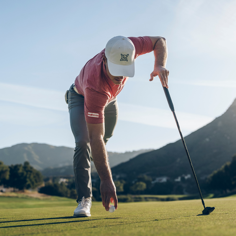 A golfer in a red shirt and white cap is placing a golf ball on the green.